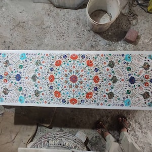 May include: A rectangular white marble table with an intricate floral inlay design. The design features a symmetrical pattern of red, blue, and green flowers and leaves. The table is a decorative piece.