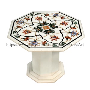Puede incluir: Mesa octogonal de mármol blanco con un diseño de incrustaciones florales. La superficie de la mesa presenta un patrón intrincado de flores y hojas en tonos de rojo, naranja, verde, azul y blanco. La mesa tiene una sola base de pedestal.