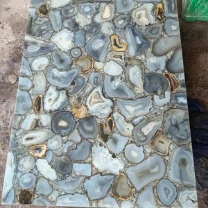 May include: A rectangular tabletop composed of irregularly shaped, polished agate stones. The stones are primarily shades of blue, gray, and white, with accents of gold and brown. The surface has a glossy finish, creating a unique and decorative element.