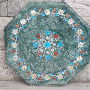 May include: An octagonal green marble table with intricate floral inlay. The table features a central floral design in blue, red, and silver, surrounded by a border of white and silver flowers with blue accents.