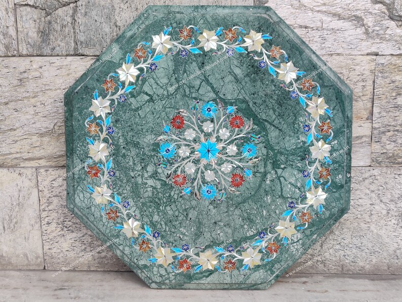 May include: An octagonal green marble table with intricate floral inlay. The table features a central floral design in blue, red, and silver, surrounded by a border of white and silver flowers with blue accents.