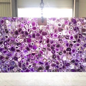 May include: Large rectangular slab of polished amethyst gemstone. The surface is a mosaic of purple, white, and brown agate slices, creating a unique pattern. The slab is suspended from a metal hook.