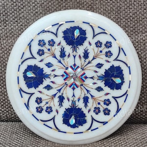 May include: Round white marble decorative piece with intricate floral inlay. The design features blue, gold, and multi-colored accents. The detailed pattern includes large stylized flowers and smaller floral elements.