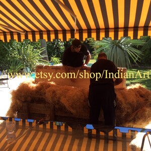 May include: Two people are working together to package a large, rectangular object wrapped in brown packing material. The object is sitting on a wooden pallet. The background shows a striped awning, green plants, and a swimming pool.