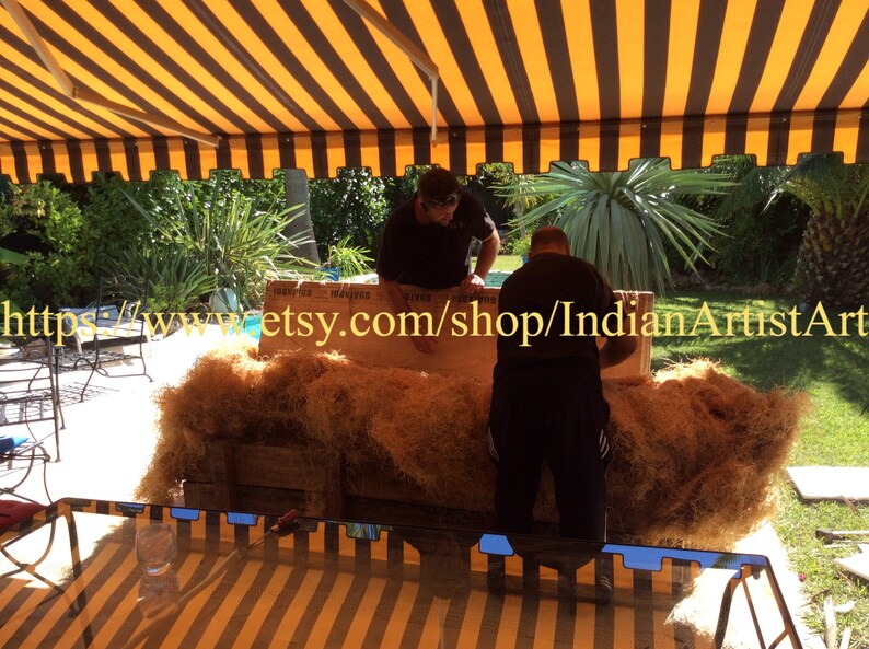 May include: Two people are working together to package a large, rectangular object wrapped in brown packing material. The object is sitting on a wooden pallet. The background shows a striped awning, green plants, and a swimming pool.