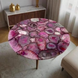 Könnte beinhalten: Ein runder Couchtisch mit einer einzigartigen Oberfläche aus rosa und lila Achat-Scheiben. Der Tisch hat eine glänzende Oberfläche und wird von drei Holzbeinen getragen. Das Achatmuster erzeugt ein auffälliges, dekoratives Element.