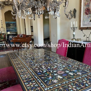 May include: A black marble dining table with intricate floral inlay work in various colors, including white, gold, blue, and green. The table is surrounded by red velvet chairs with a black and white patterned design. A large crystal chandelier hangs above the table.