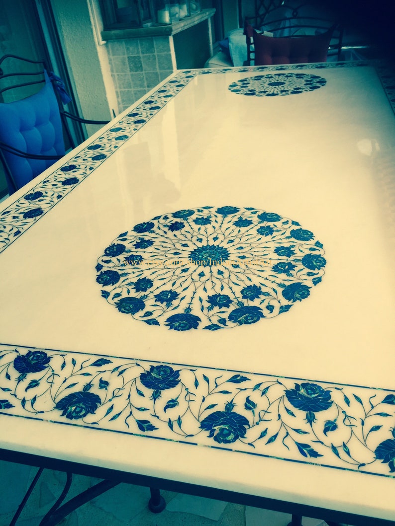 May include: A white marble table top with intricate blue floral designs inlaid into the surface. The table top is polished and reflects light.