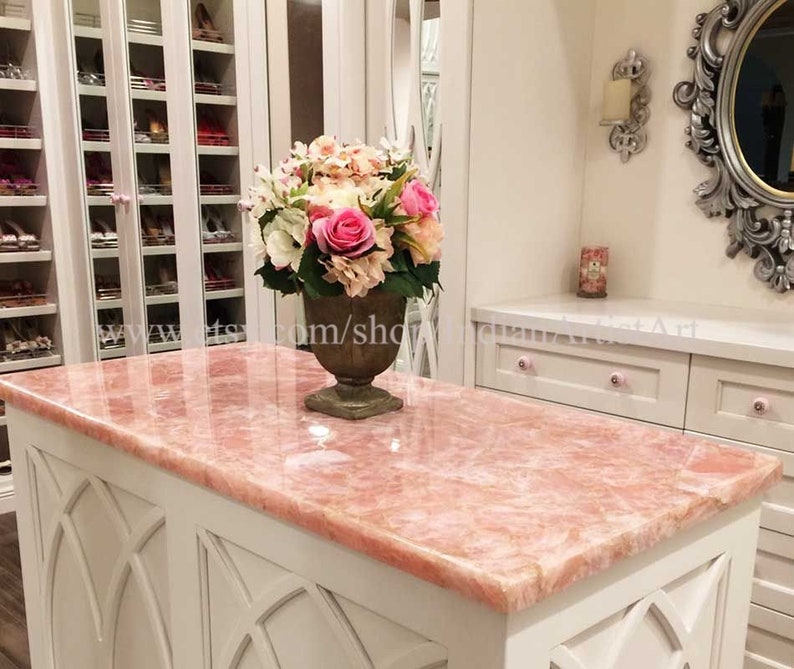 May include: A pink marble countertop with a white base and ornate detailing. A bouquet of pink and white flowers in a brown ceramic vase sits on the countertop.