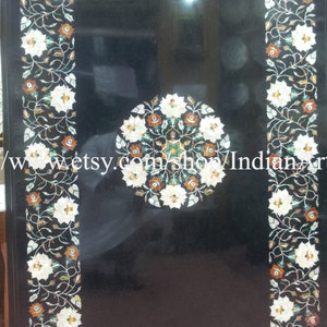 Puede incluir: Tapa de mesa de mármol negro con un diseño floral incrustado con piedras blancas, verdes y naranjas. El diseño presenta un círculo central con una estrella en el medio.