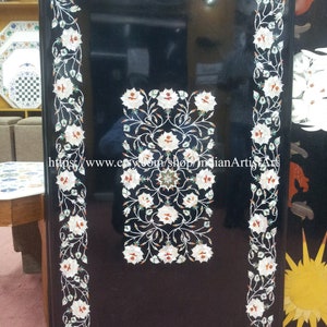 May include: A rectangular black marble table top with white floral inlay. The border and central design feature white flowers with red and green accents. The table top is a decorative piece.
