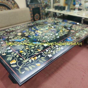 May include: A black marble table top with a floral design inlaid with colourful stones. The table top is surrounded by a black border. The table is in a room with a sofa and other furniture.