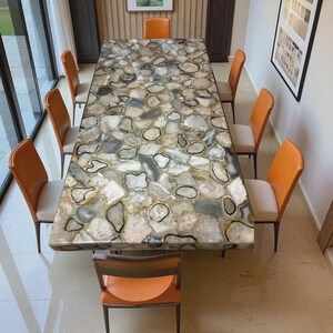May include: A large rectangular dining table with a stone top featuring a mosaic of gray, white, and black agate slices. Eight orange leather chairs with beige seats surround the table. The table is approximately 100 inches long.