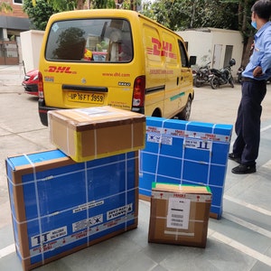 May include: A yellow DHL delivery van with a stack of four packages in front of it. The packages are wrapped in brown paper and blue tape. The top package is a brown cardboard box with a white label that says "FRAGILE HANDLE WITH CARE". The other packages are wrapped in blue paper with white tape and have "FRAGILE" labels.