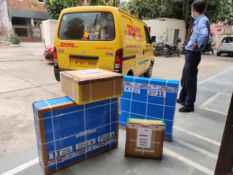 May include: A yellow DHL delivery van with a stack of four packages in front of it. The packages are wrapped in brown paper and blue tape. The top package is a brown cardboard box with a white label that says "FRAGILE HANDLE WITH CARE". The other packages are wrapped in blue paper with white tape and have "FRAGILE" labels.