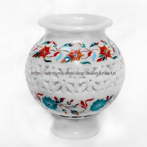 May include: White marble vase with intricate floral inlay in red, teal, and orange. The vase has a round body with a flared rim and a pedestal base. The body features a cut-out design. Text on the vase reads: https://www.etsy.com/shop/IndianArtistArt