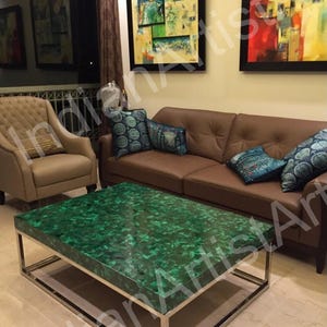 Op de afbeelding: Een groene malachiet salontafel met een zilveren metalen frame. De tafel staat in een woonkamer met een bruine leren bank en een beige fauteuil.