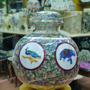 Pode incluir: Um jarro de cerâmica branca com tampa, decorado com incrustações de madrepérola e pedras coloridas. O jarro apresenta dois desenhos circulares, um com um pavão e o outro com um elefante.