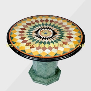 Pode incluir: Mesa redonda de mármore com tampo em mosaico com padrão geométrico em tons de amarelo, castanho, verde e branco. A mesa tem uma borda preta e uma base octogonal verde. A mesa é uma peça decorativa.