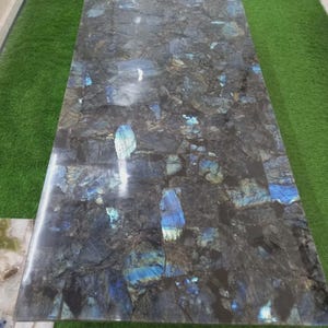 May include: A rectangular table with a polished surface, showcasing a mosaic of dark gray, black, and blue-green stone fragments. The stone has iridescent flashes of blue and green. The table is set against a green background.