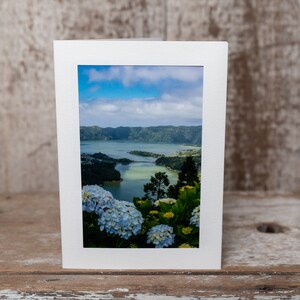 May include: A white card with a photo of a lake surrounded by green hills and blue sky. The lake is a light blue color and there are white flowers in the foreground.