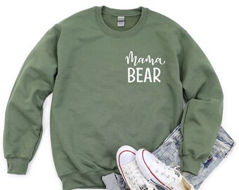May include: A green sweatshirt with the text "Mama Bear" in white lettering on the front. The sweatshirt is shown on a white background with a pair of white sneakers and blue jeans.