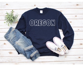 Può includere: Una felpa blu navy con la scritta "OREGON" stampata in bianco sul davanti. La felpa è abbinata a jeans blu e scarpe da ginnastica bianche.