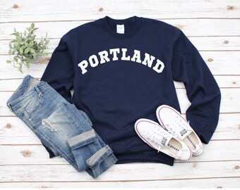 May include: A navy blue sweatshirt with the word "PORTLAND" printed in white on the front. The sweatshirt is paired with blue jeans and white trainers.