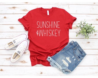 May include: Red t-shirt with white text that says "Sunshine & Whiskey". The shirt is laid flat on a white wooden surface with a pair of white sneakers, denim shorts, and a small potted plant.
