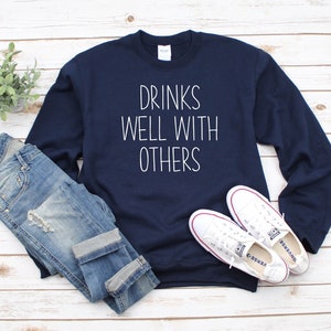 Może przedstawiać: Granatowa bluza z białym napisem "Drinks Well With Others". Bluza jest zestawiona z niebieskimi jeansami i białymi butami sportowymi.