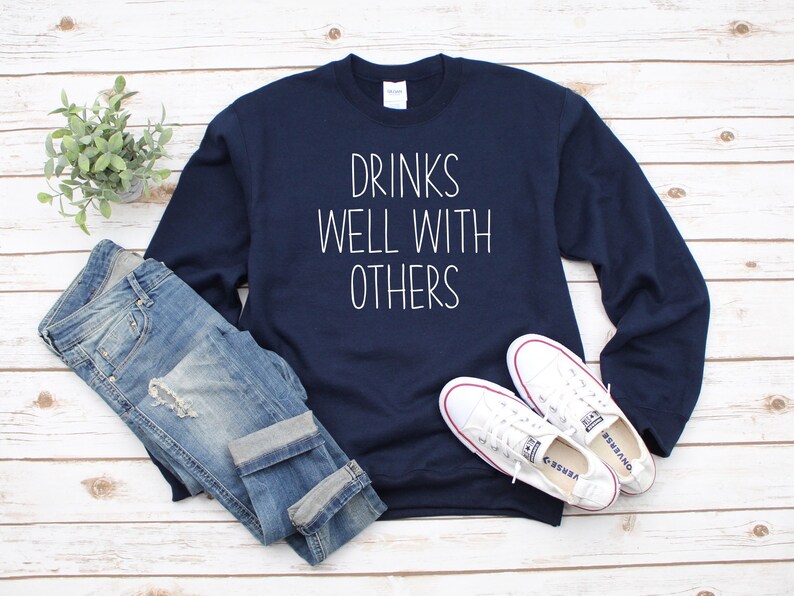 Może przedstawiać: Granatowa bluza z białym napisem "Drinks Well With Others". Bluza jest zestawiona z niebieskimi jeansami i białymi butami sportowymi.