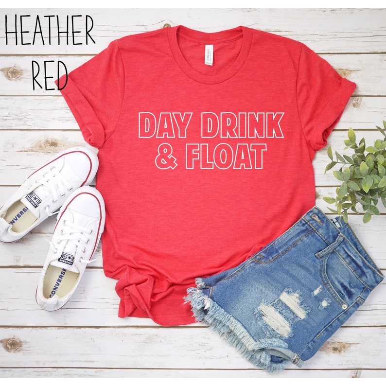 Op de afbeelding: Rood heather t-shirt met witte tekst die "DAY DRINK & FLOAT" luidt. Het shirt ligt plat op een wit houten oppervlak met een paar witte canvas schoenen en een spijkerbroek.