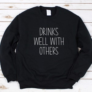 Może przedstawiać: Czarna bluza z białym napisem "Drinks well with others".
