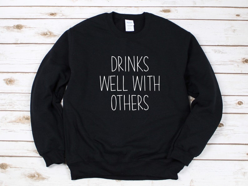 Może przedstawiać: Czarna bluza z białym napisem "Drinks well with others".