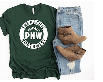 Może przedstawiać: Zielony t-shirt z białym kołem z napisem "The Pacific Northwest" i górami w środku.