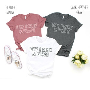 Op de afbeelding: Drie T-shirts met de tekst "Day Drink & Float" in witte omrande letters. De shirts zijn in heide-mauve, wit en donker heide grijs.