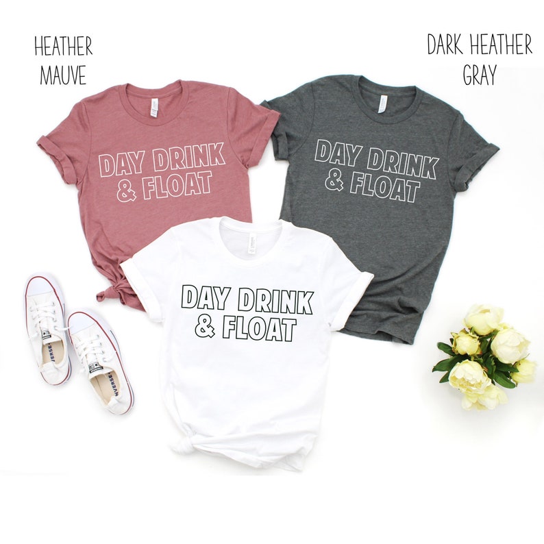 Op de afbeelding: Drie T-shirts met de tekst "Day Drink & Float" in witte omrande letters. De shirts zijn in heide-mauve, wit en donker heide grijs.