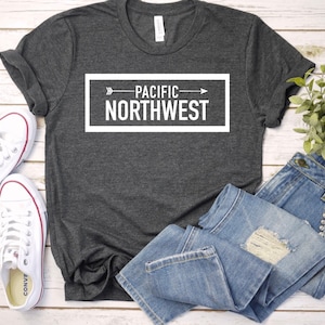 May include: A dark gray t-shirt with a white graphic that says "PACIFIC NORTHWEST" with an arrow pointing to the right. The shirt is laid out on a wooden surface with a pair of white sneakers and blue jeans.
