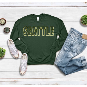 Peut inclure: Un sweat-shirt vert fonc&eacute; avec le mot "SEATTLE" imprim&eacute; en lettres jaunes soulign&eacute;es.