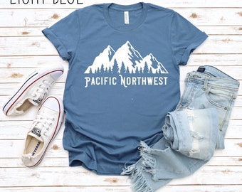 Może przedstawiać: Jasnoniebieska koszulka z białym nadrukiem gór i drzew. Pod nadrukiem znajduje się napis "Pacific Northwest".