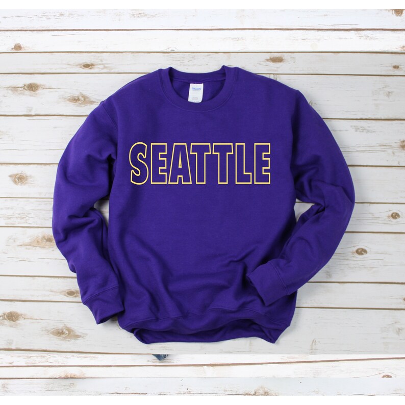 Peut inclure: Sweat-shirt violet avec un contour jaune du mot "SEATTLE" sur le devant.