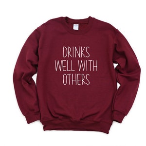 Może przedstawiać: Bordowa bluza z napisem "Drinks Well With Others" w kolorze białym.