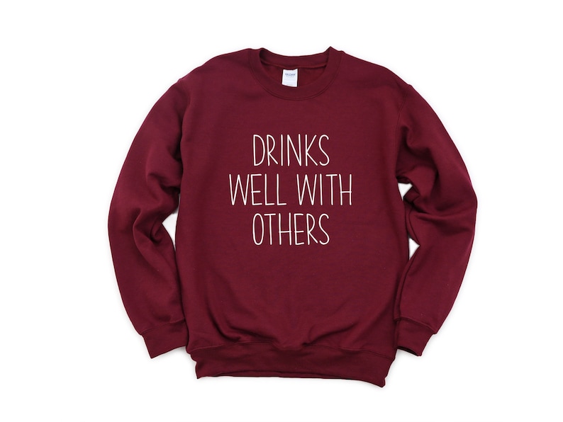 Może przedstawiać: Bordowa bluza z napisem "Drinks Well With Others" w kolorze białym.