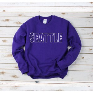 Peut inclure: Sweat-shirt violet avec un texte blanc encadr&eacute; qui dit "SEATTLE".