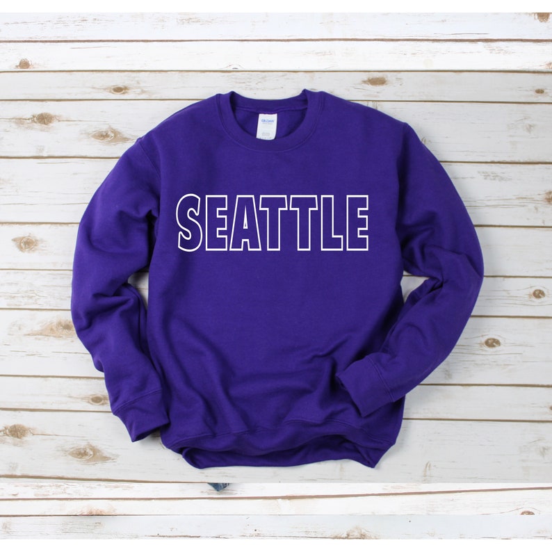 Peut inclure: Sweat-shirt violet avec un texte blanc encadr&eacute; qui dit "SEATTLE".