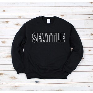 Peut inclure: Sweat-shirt noir avec un texte blanc qui dit "SEATTLE" sur le devant.