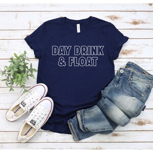 Op de afbeelding: Een marineblauw t-shirt met witte tekst die "DAY DRINK & FLOAT" zegt. Het shirt ligt op een wit houten oppervlak met een paar witte sneakers en een blauwe jeans.
