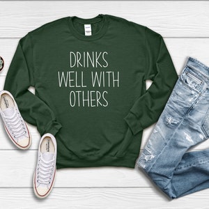Może przedstawiać: Zielony sweter z napisem "Drinks Well With Others" w białym kolorze.