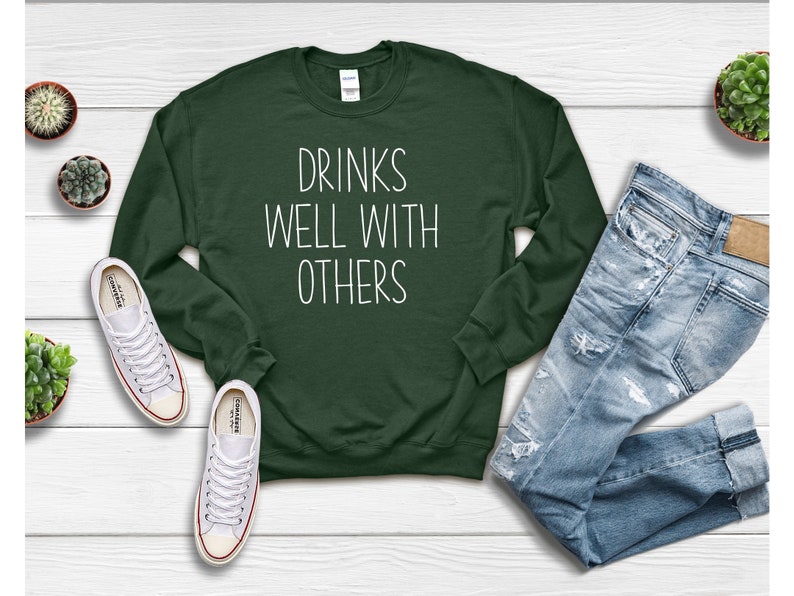 Może przedstawiać: Zielony sweter z napisem "Drinks Well With Others" w białym kolorze.