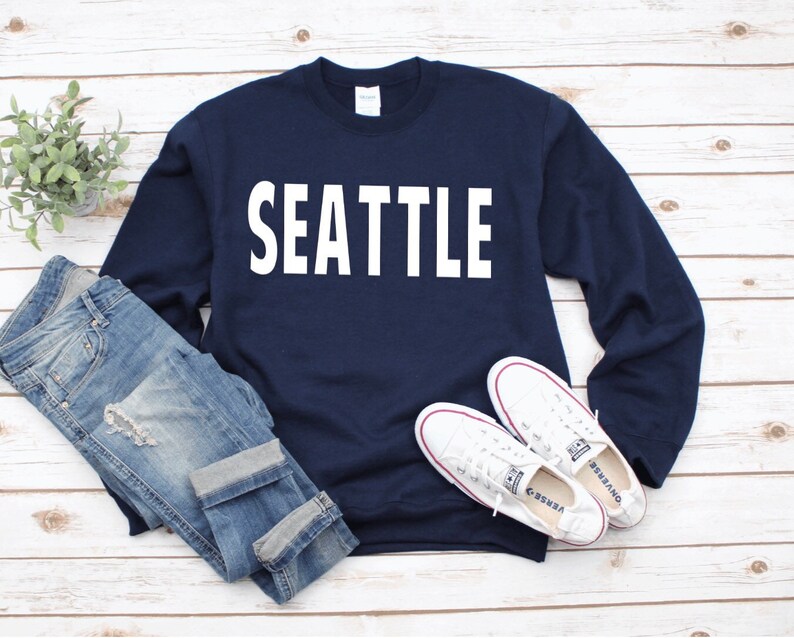 May include: A navy blue sweatshirt with the word "SEATTLE" printed in white on the front. The sweatshirt is laid out on a white wooden surface with a pair of white trainers and blue jeans.